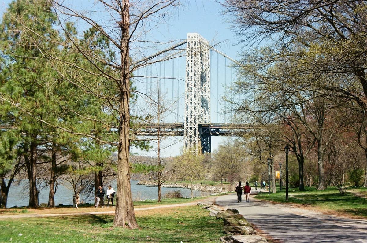 Parkszene am Flussufer mit Spaziergängern, Bäumen und der George Washington Bridge im Hintergrund in Upper Manhattan.