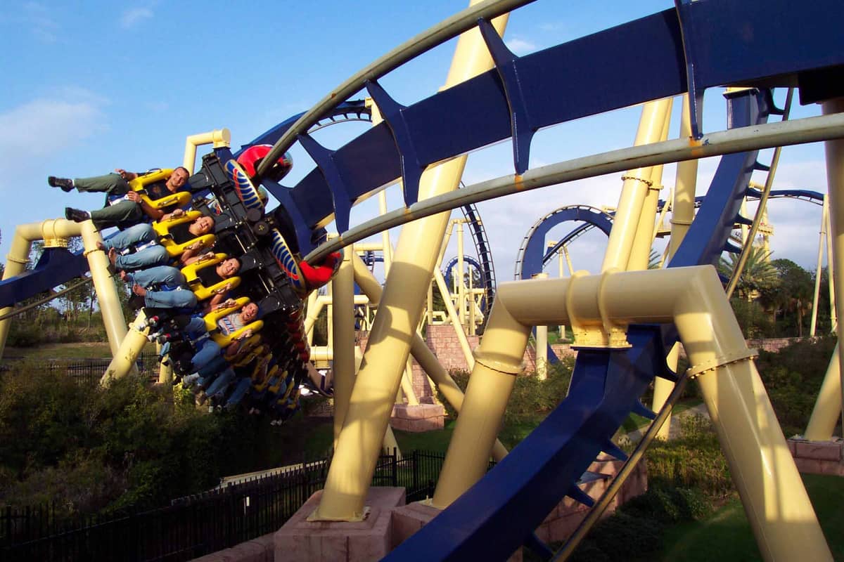 Menschen fahren eine Looping-Achterbahn mit gelber und blauer Schiene in einem Freizeitpark, umgeben von Grün und klarem Himmel.