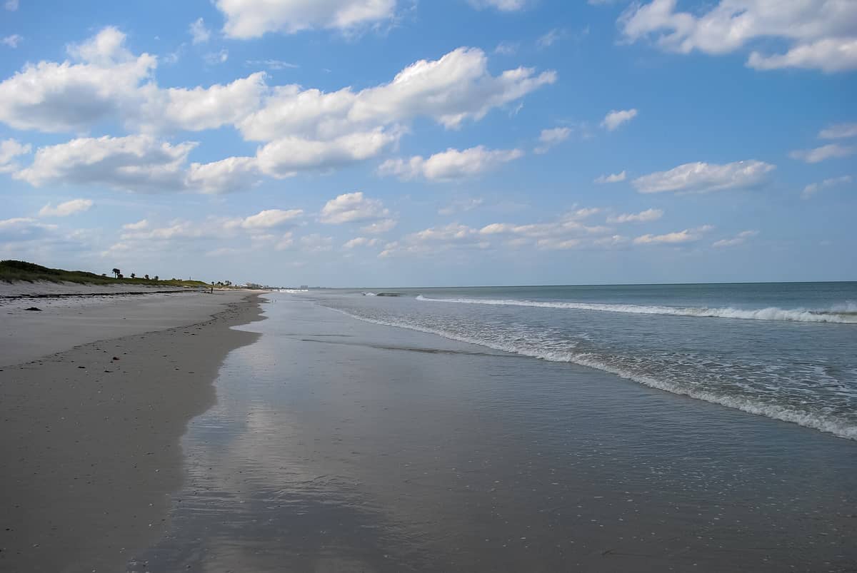 Breiter Sandstrand mit sanften Wellen unter einem blauen, leicht bewölkten Himmel, der einen ruhigen Abschnitt der Atlantikküste Floridas zeigt.