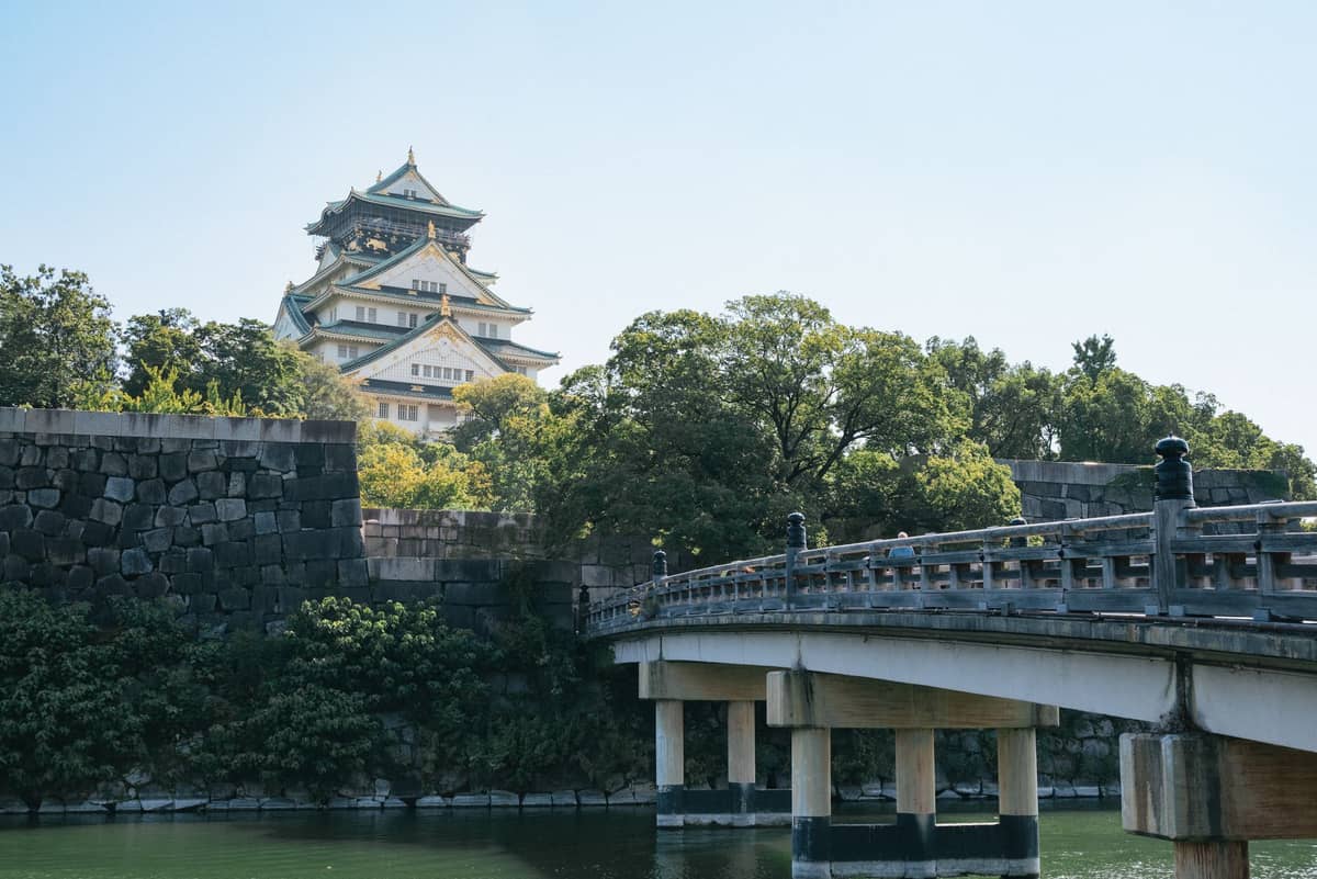 Osaka Castle with its stone walls and a wooden bridge crossing the moat, surrounded by trees under a clear blue sky.