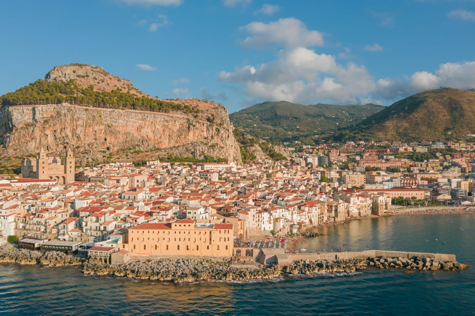 Luftaufnahme der sizilianischen Küstenstadt Cefalù mit mittelalterlichen Gebäuden, historischer Kathedrale und dramatischen Felsklippen am Tyrrhenischen Meer unter klarem Himmel.