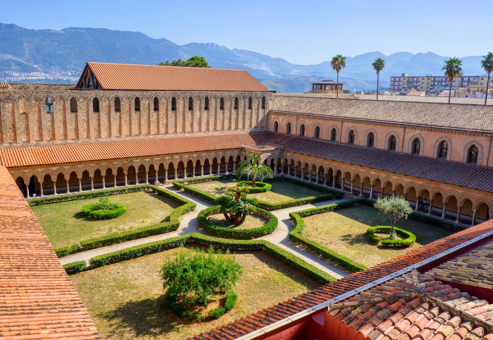 Luftaufnahme des Kreuzgangs der Kathedrale von Monreale mit kunstvollen Säulen, Gartenwegen und einer fernen Gebirgslandschaft unter einem klaren blauen Himmel.