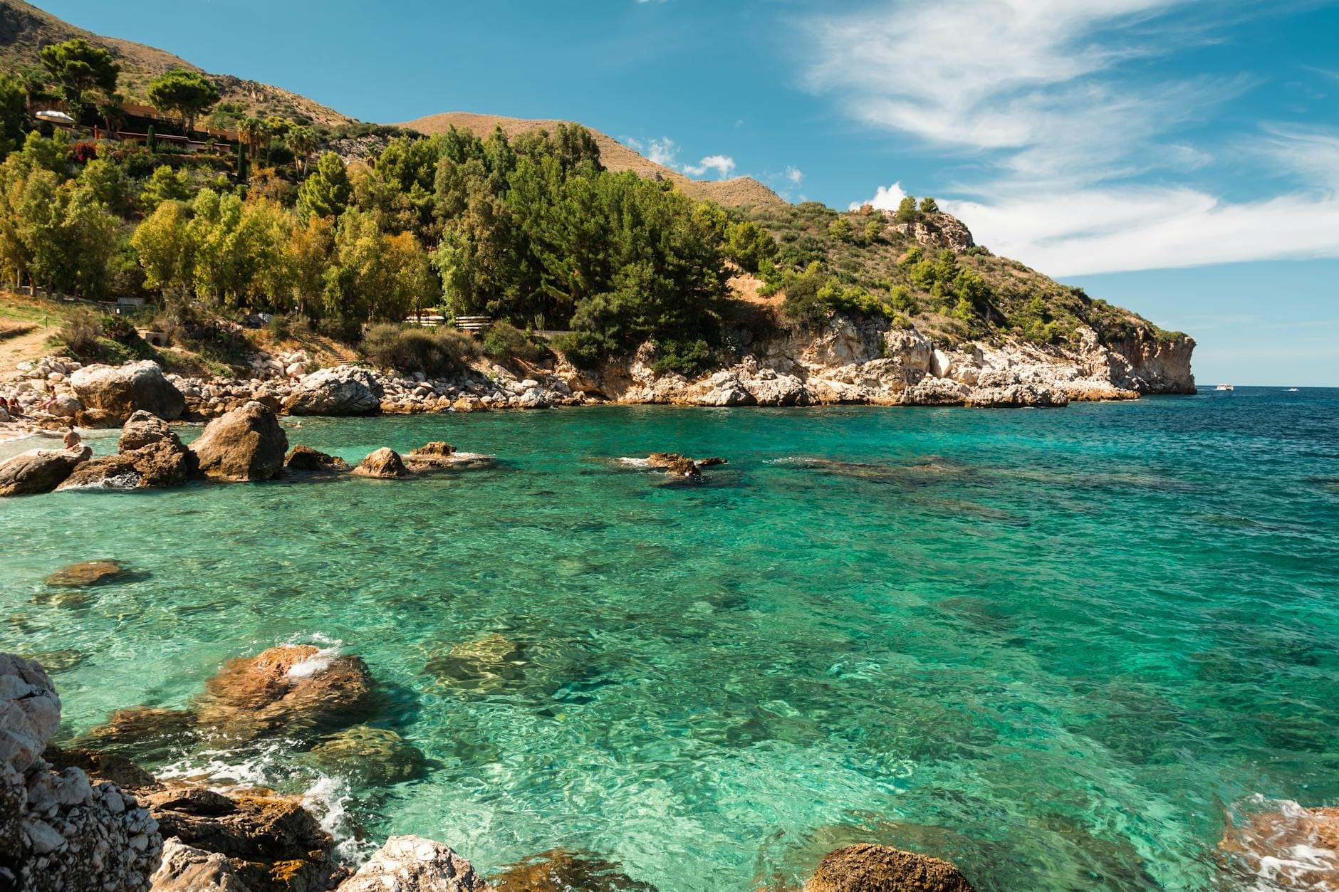Felsige Küste mit kristallklarem türkisfarbenem Wasser und üppigen grünen Bäumen unter einem blauen Himmel, typisch für die Naturschutzgebiete Siziliens.