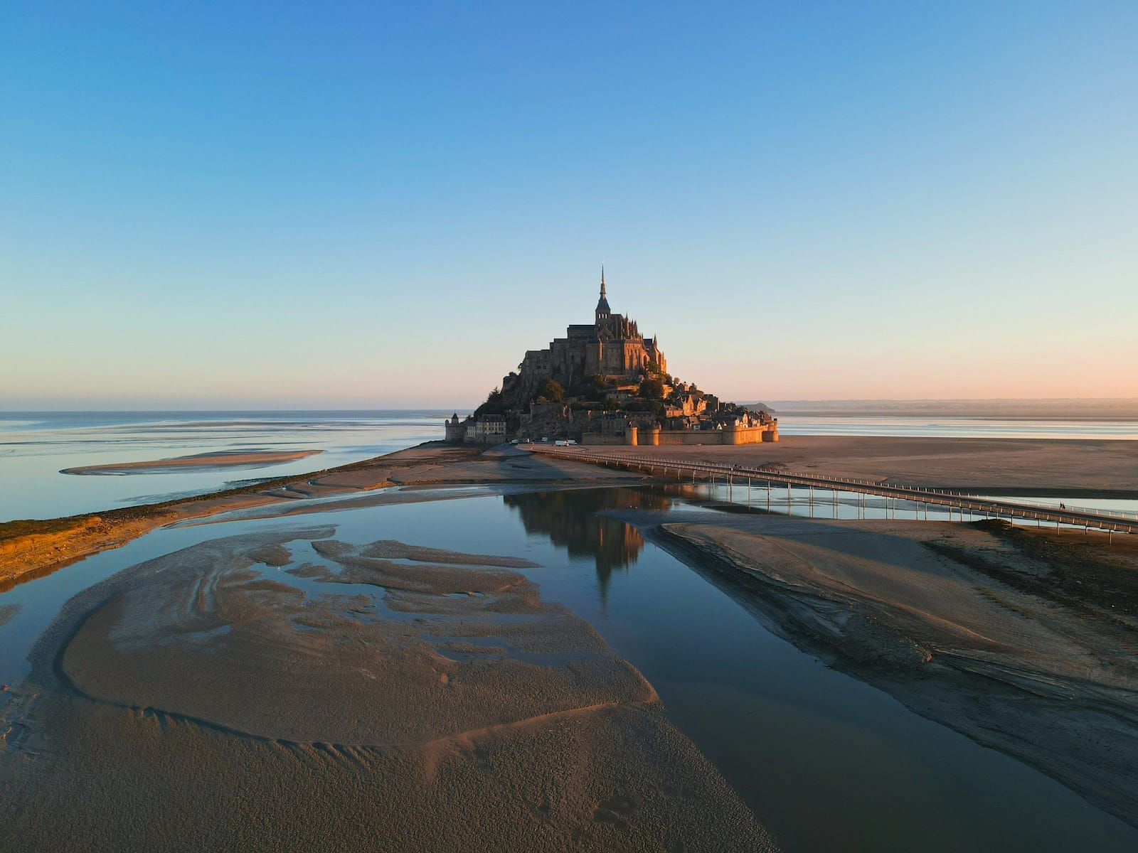 Mont Saint-Michel'in gün doğumunda gelgit sularındaki yansımaları ve açık mavi gökyüzüyle uzaktan görünümü.