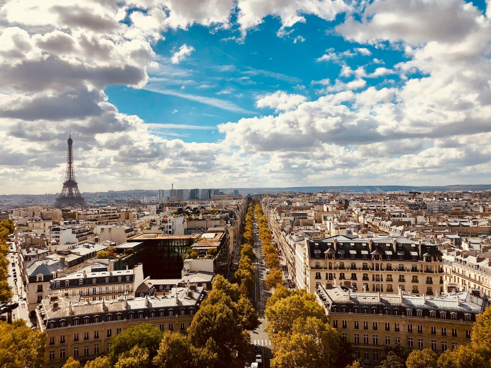 Vista panoramica di Parigi con la Torre Eiffel a sinistra, viali alberati e un cielo azzurro con nuvole sparse.
