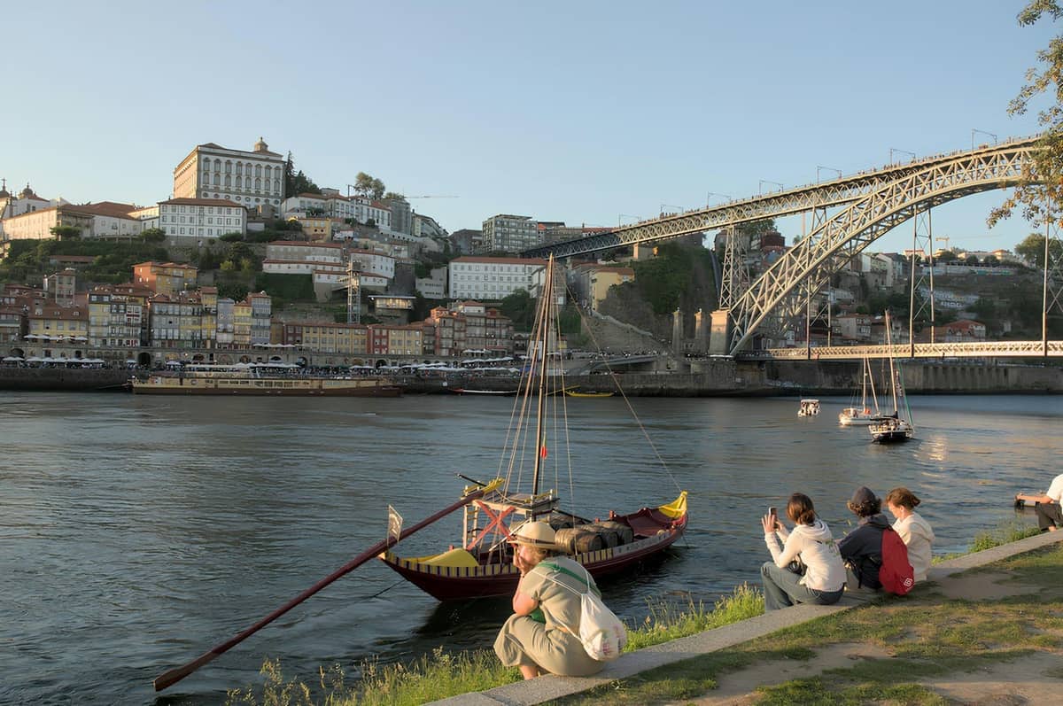 Menschen, die sich am Flussufer in Vila Nova de Gaia entspannen, mit traditionellen Booten auf dem Douro und der Brücke Dom Luís I, die nach Porto führt