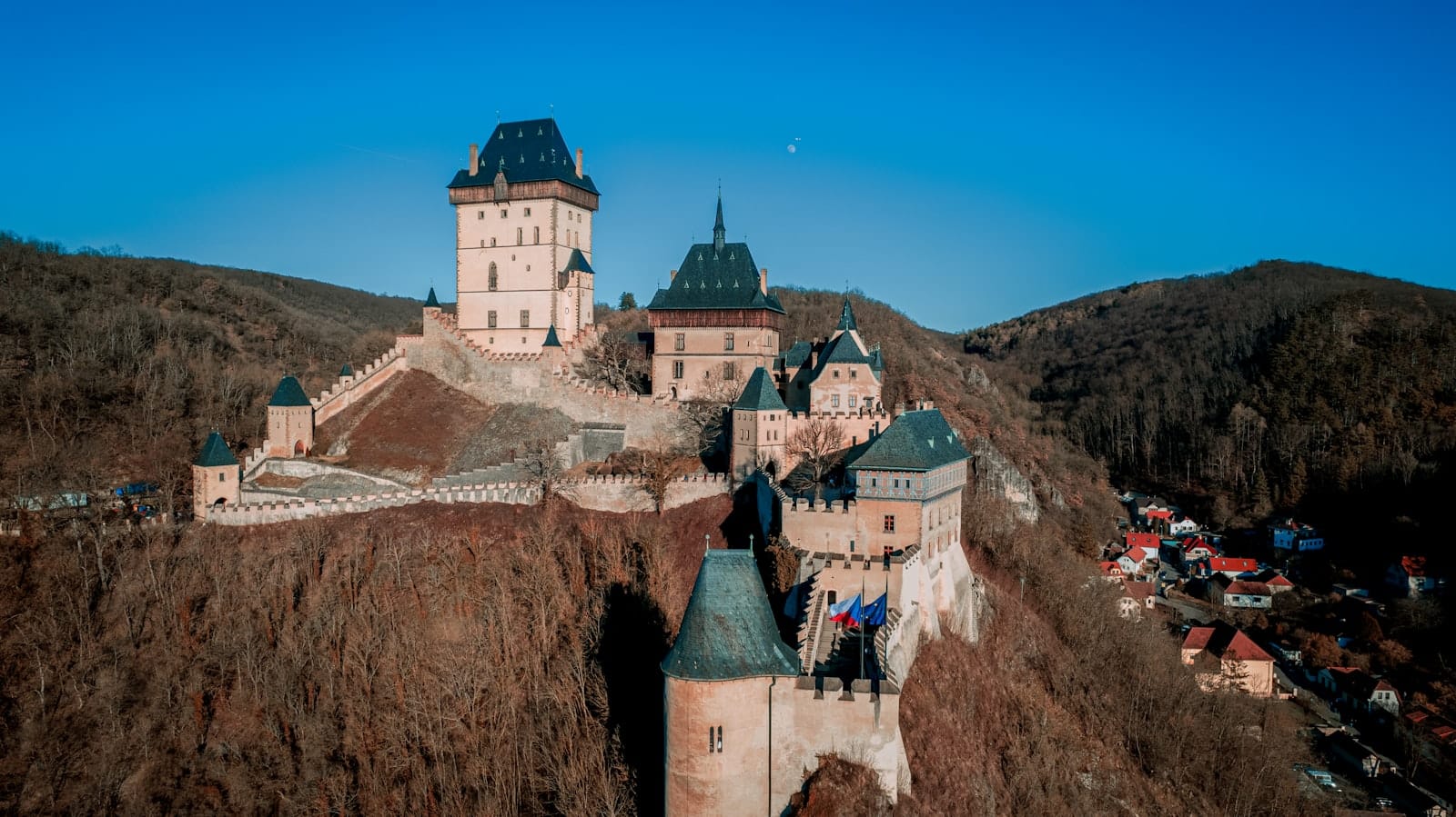 Vista aerea del Castello di Karlštejn arroccato su una collina boscosa con il villaggio sottostante, sotto un cielo azzurro limpido