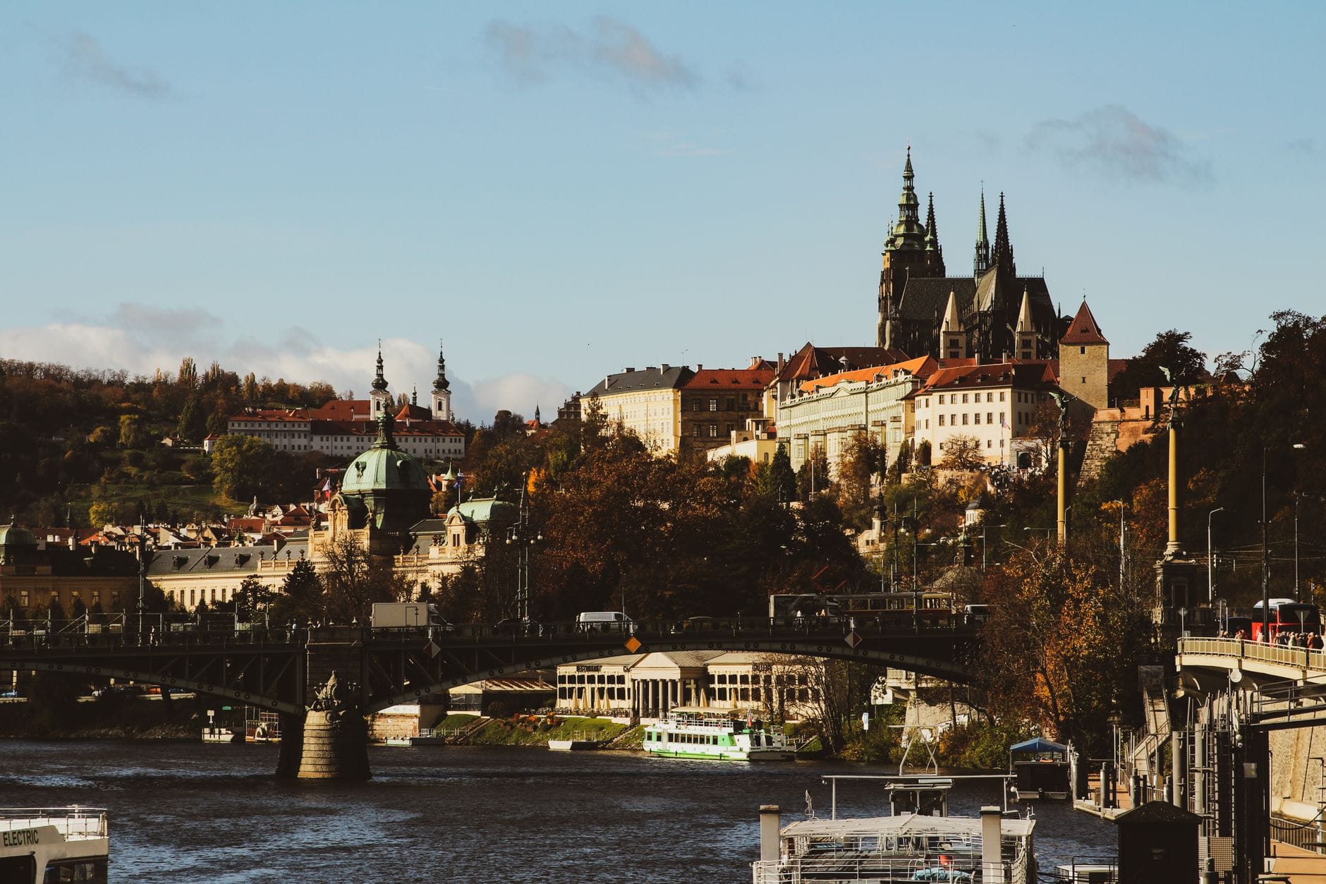 Vista diurna del Castello di Praga, tetti rossi e il fiume Vltava con ponti e vivace attività cittadina in primo piano.