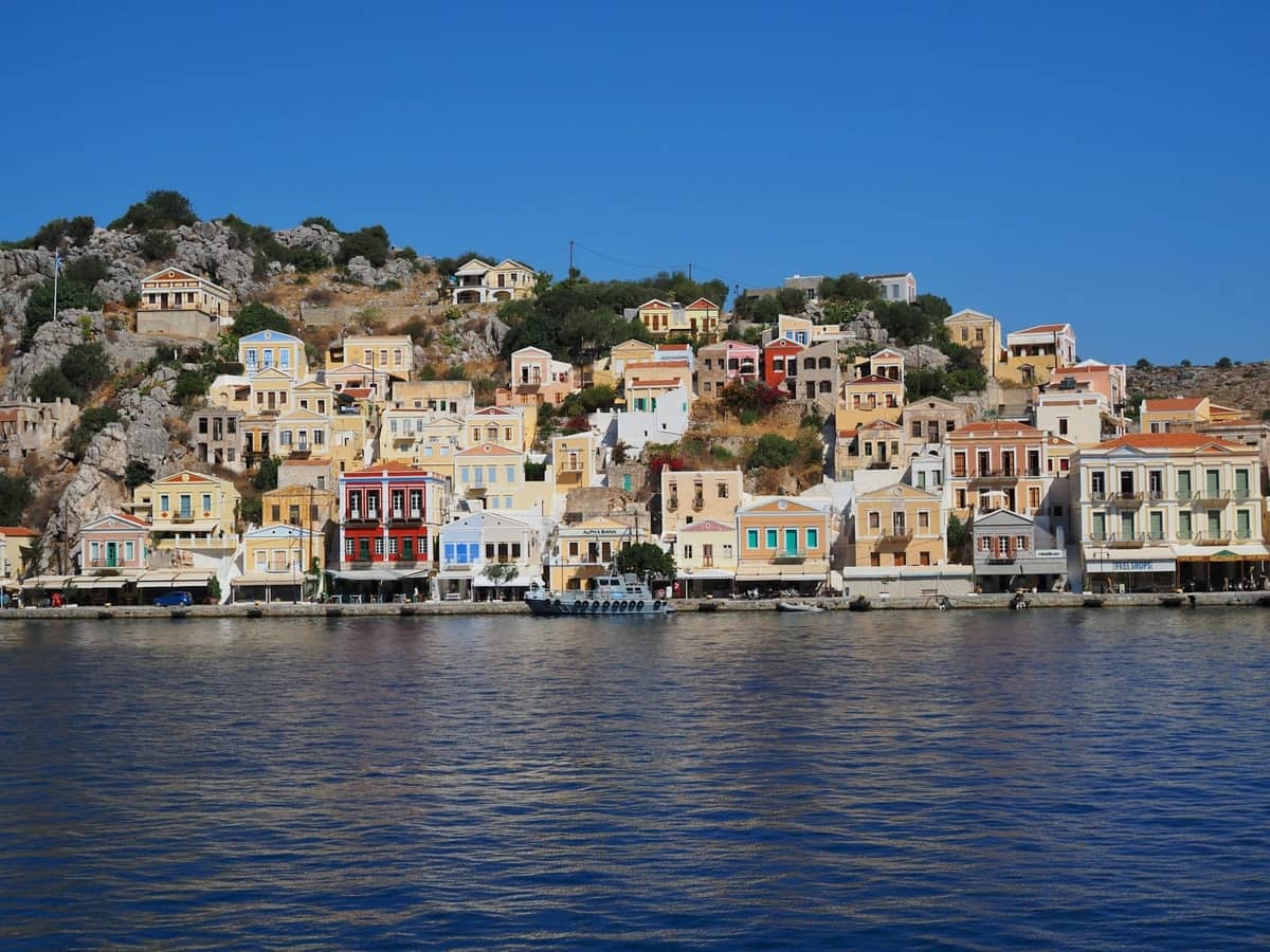 Pastel-colored neoclassical houses rising up a hillside above blue water, typical of Symi island, a popular ferry day trip from Rhodes