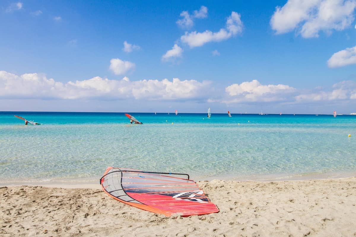 Sandy beach in southern Rhodes with clear turquoise water, windsurf boards and windsurfers in the distance under a blue sky.