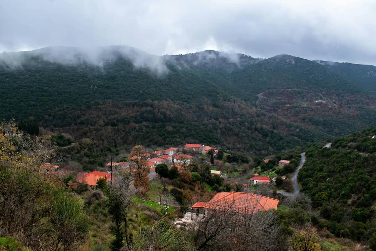 Small village of red-roofed houses nestled in the green, misty mountains of Rhodes’ interior, with a winding road and forested hills.