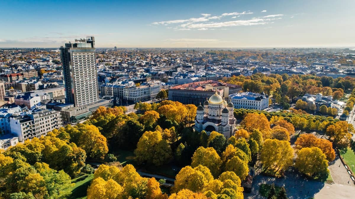 Luftaufnahme der Stadt Riga mit den goldenen Kuppeln der Christi-Geburts-Kathedrale, umgeben von grünen und gelben Herbstbäumen unter klarem blauem Himmel.