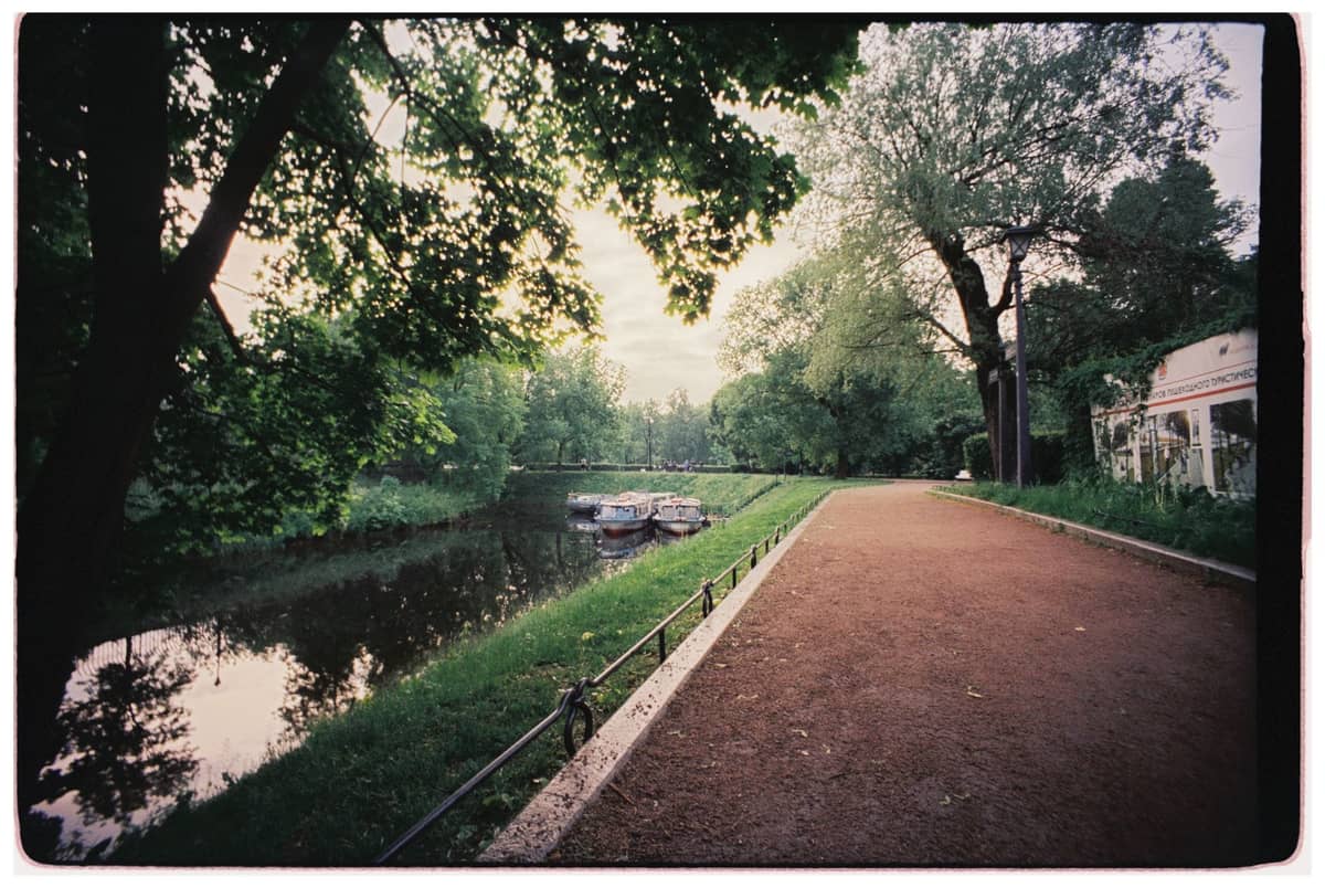 Grüner Parkweg am Flussufer mit mehreren festgemachten kleinen Booten, Bäume wölben sich über das Wasser, ruhige Umgebung nahe dem Stadtzentrum.