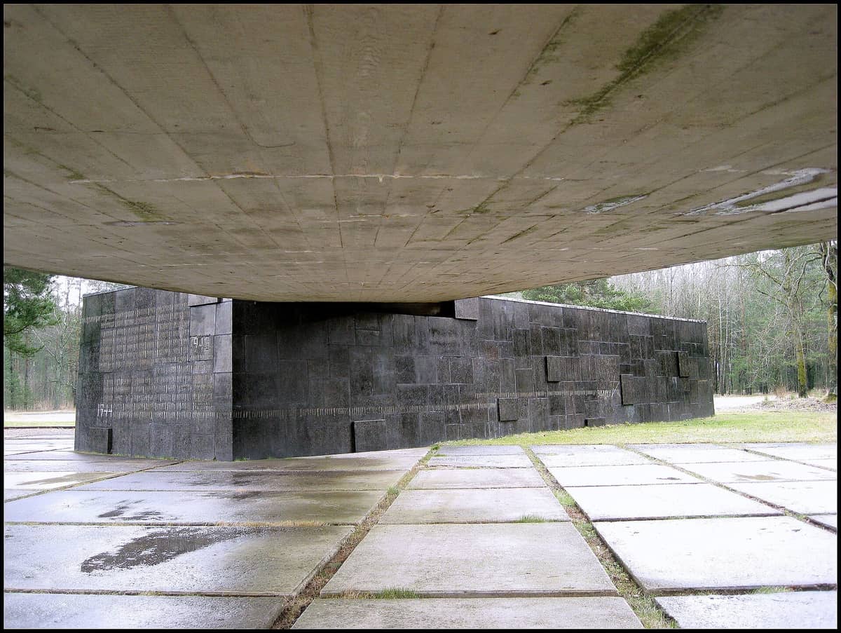Blick auf ein riesiges Betondenkmal mit schwarzen Steinmauern am Salaspils Memorial, umgeben von Wald und nüchternem Pflaster.