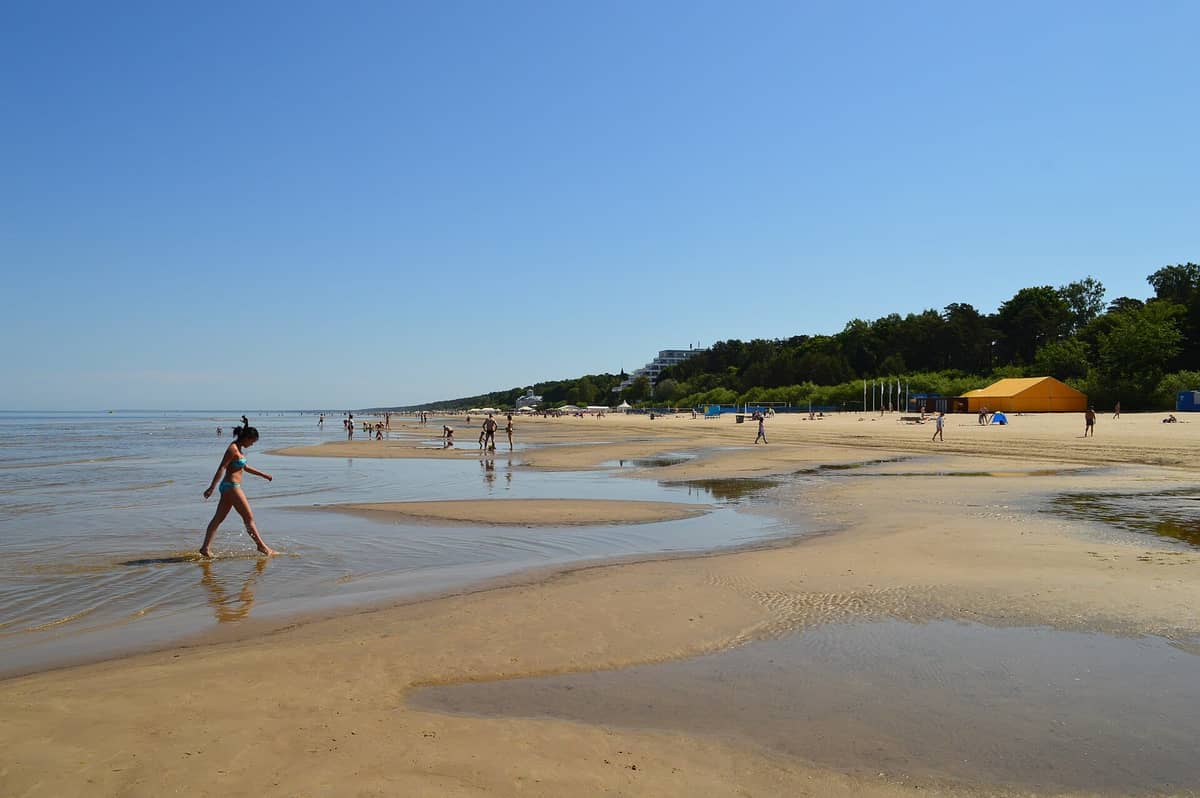 Sonniger Sommertag am Strand von Jūrmala, mit Menschen, die schwimmen, auf nassem Sand spazieren gehen und Kiefernwäldern im Hintergrund.