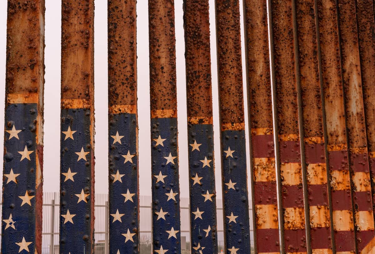 Nahaufnahme des Grenzzauns zwischen den USA und Mexiko mit verrosteten, mit der amerikanischen Flagge bemalten Stäben im Freien bei Tageslicht.
