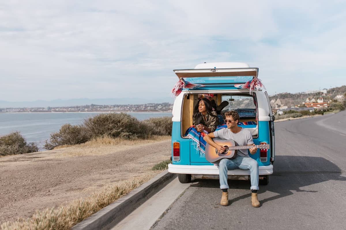 Zwei Personen sitzen am Heck eines blauen Vans, der auf einer Küstenstraße nahe San Diego parkt – im Hintergrund Meer und Stadtsilhouette.