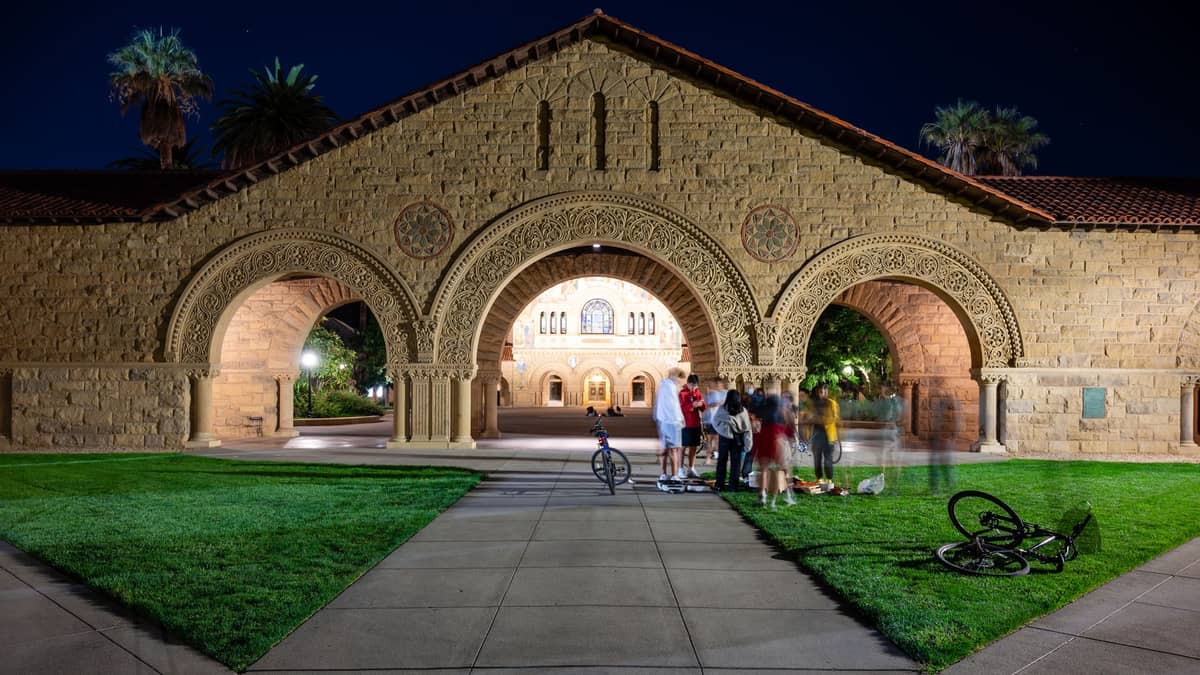 Vista notturna del caratteristico porticato ad archi della Stanford University con un gruppo di persone sul prato e biciclette in primo piano.