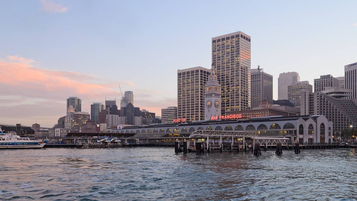 Il Ferry Building di San Francisco e il lungomare visti dalla baia al tramonto, con traghetti, moli e lo skyline del centro città.