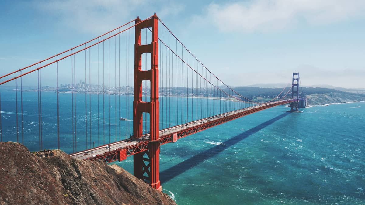 Vista aérea panorámica del puente Golden Gate sobre las aguas azules de la bahía, con las colinas de Marin y el horizonte de San Francisco al fondo en un día despejado.