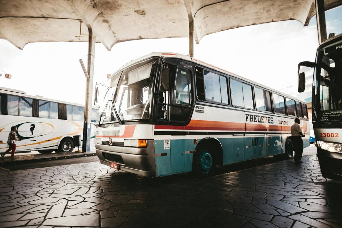 Ein klassischer blau-weißer Bus, der an einem überdachten Terminal geparkt ist, mit Fahrgästen in der Nähe, was auf den typischen öffentlichen Nahverkehr der griechischen Inseln hindeutet.