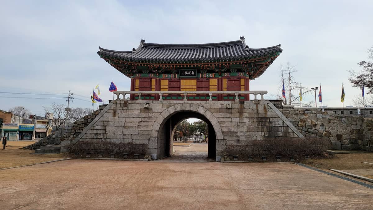 Vista em grande angular de um histórico portão de fortaleza coreana com telhado colorido e entrada em arco de pedra, bandeiras e praça aberta à frente.