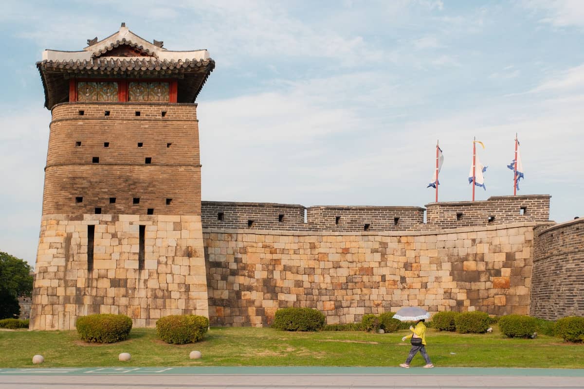 Vista ampla das muralhas de pedra e da torre da Fortaleza Suwon Hwaseong com bandeiras e uma pessoa caminhando em frente.