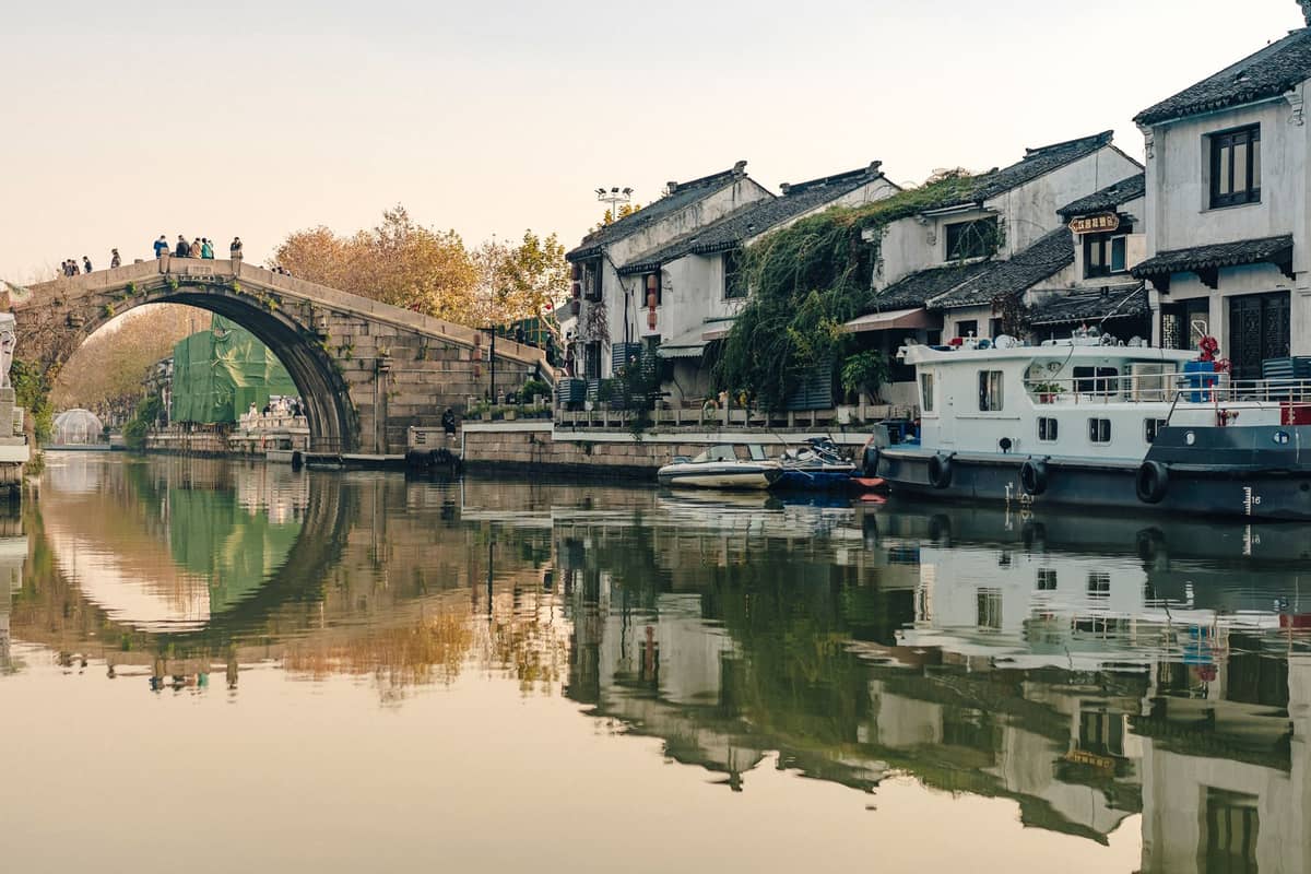 Puente de piedra con arcos sobre un canal con casas encaladas históricas y barcas, reflejando el paisaje clásico del Pueblo Acuático de Zhujiajiao en un día tranquilo.