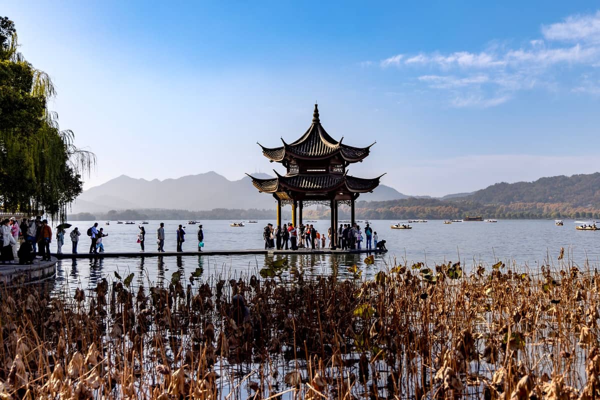 Personas pasean por un pabellón a orillas del lago con arquitectura tradicional china en el Lago del Oeste, Hangzhou, con montañas al fondo.