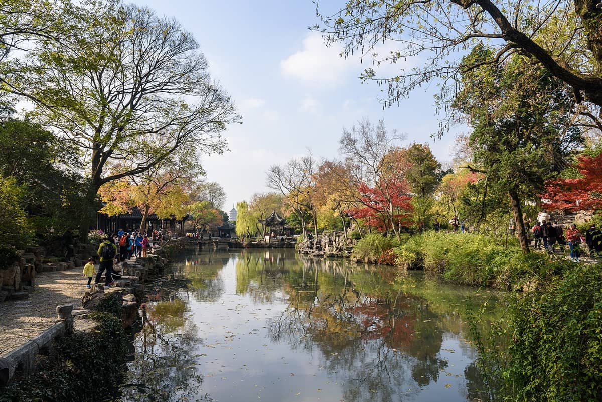 Personas paseando a orillas de un estanque arbolado con pabellones tradicionales en un jardín clásico chino en Suzhou.