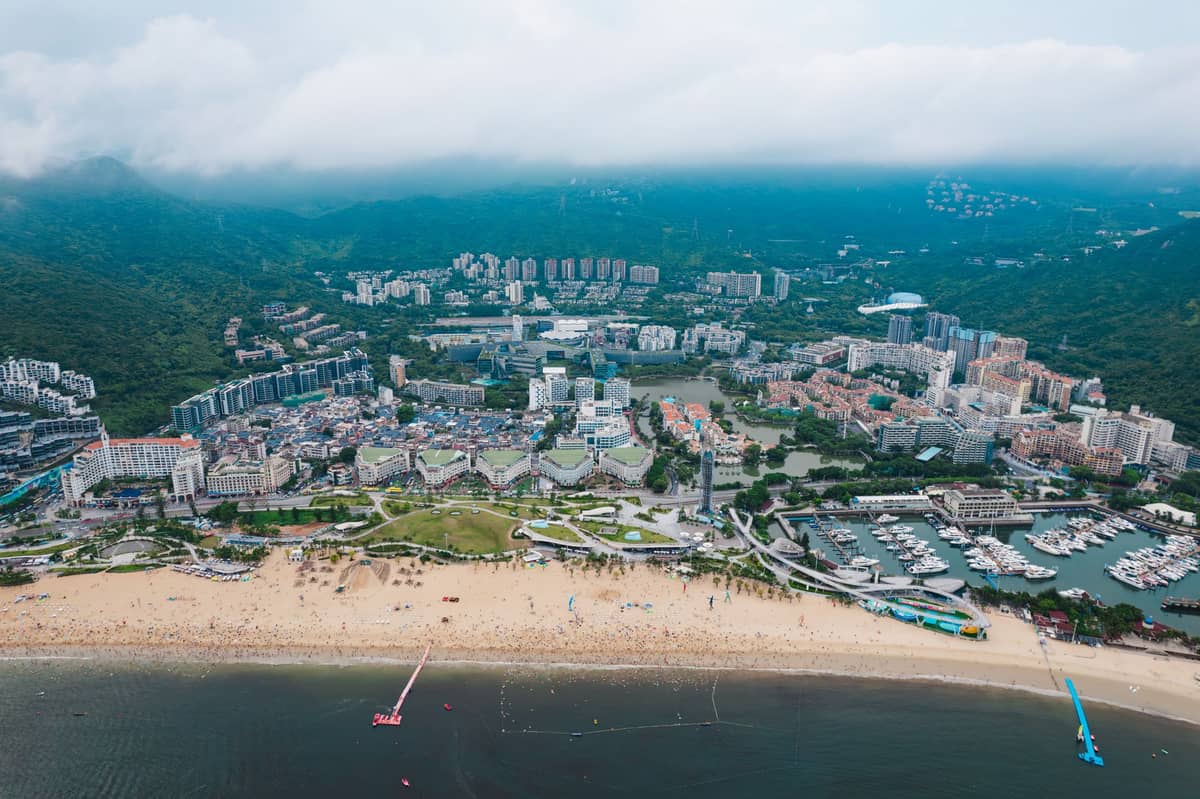Vista aérea de uma praia de areia, marina e cidade costeira com colinas verdes ao fundo em Dapeng, Shenzhen, sob céus nublados.
