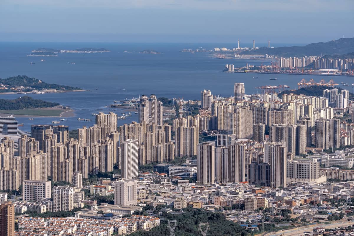 Uma ampla vista aérea de Shenzhen com densos edifícios de grande altura, colinas ao redor e a linha costeira, ilustrando a localização estratégica da cidade.