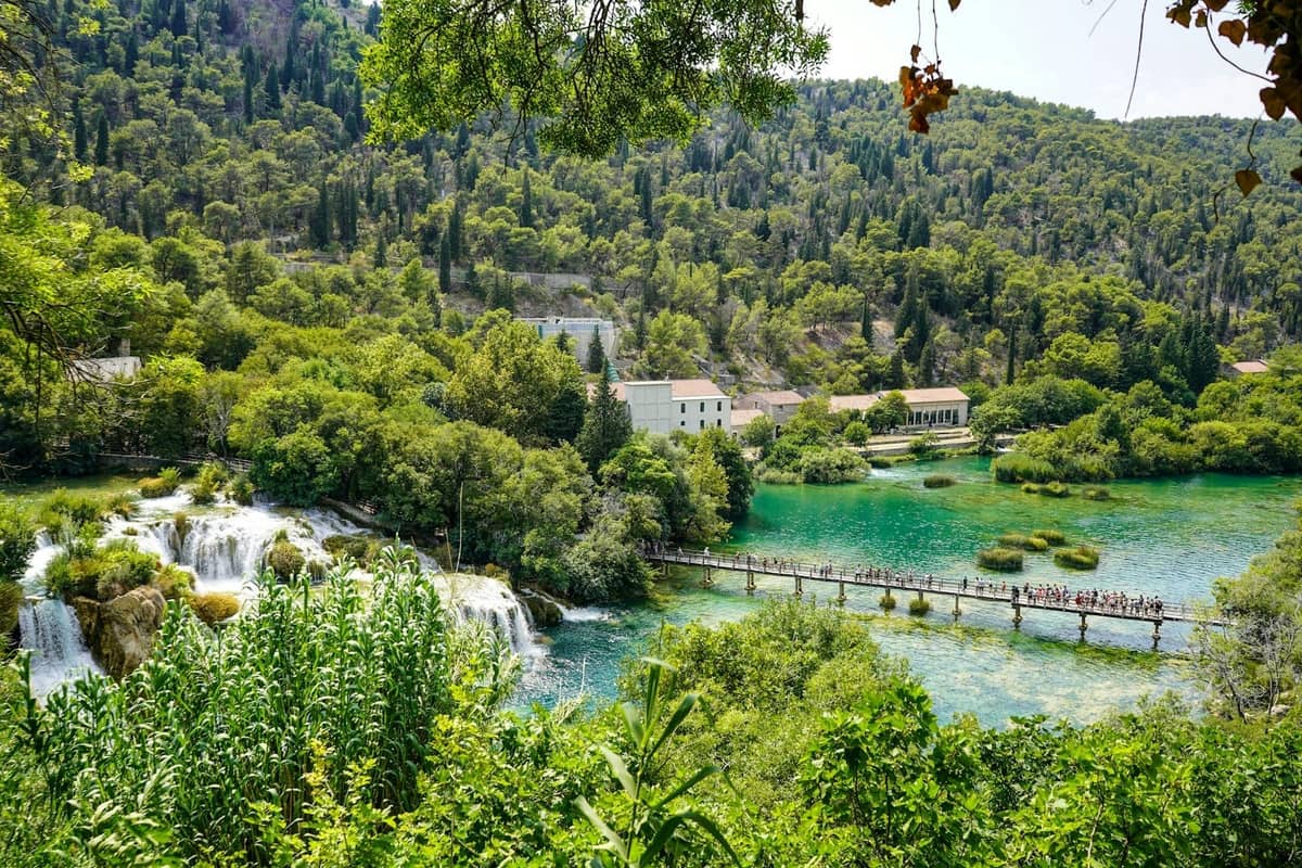 Blick auf den Nationalpark Krka mit einem Wasserfall, klarem türkisfarbenem Wasser, einer Fußgängerbrücke mit Besuchern, umgeben von üppigen grünen Hügeln.