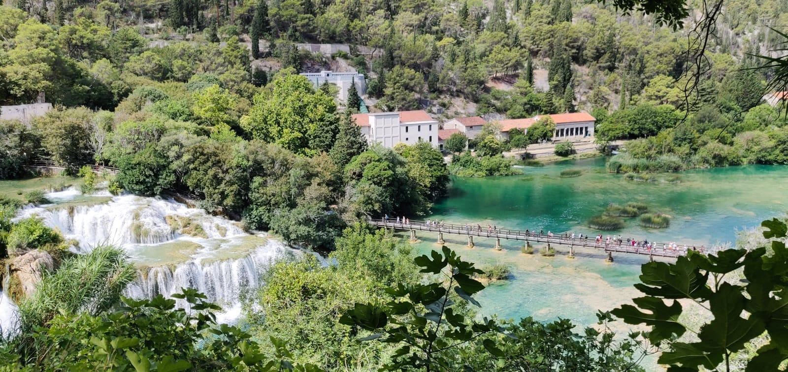 Vista panorâmica do Parque Nacional de Krka com as famosas cachoeiras Skradinski Buk, rio turquesa, construções históricas de pedra e uma passarela de madeira cercada por vegetação exuberante.