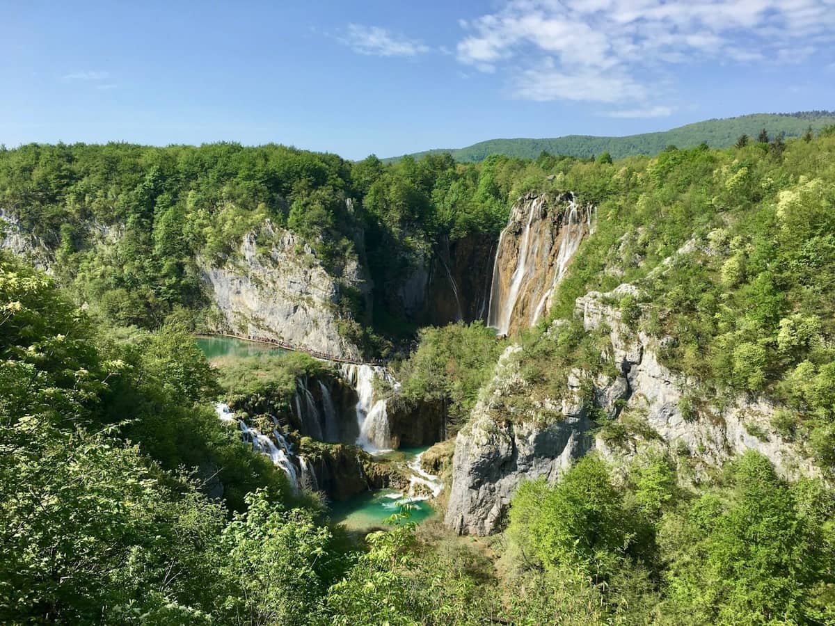 Vista de las impresionantes cascadas y piscinas turquesas de los Lagos de Plitvice rodeadas de exuberante bosque verde y acantilados rocosos bajo un cielo azul.