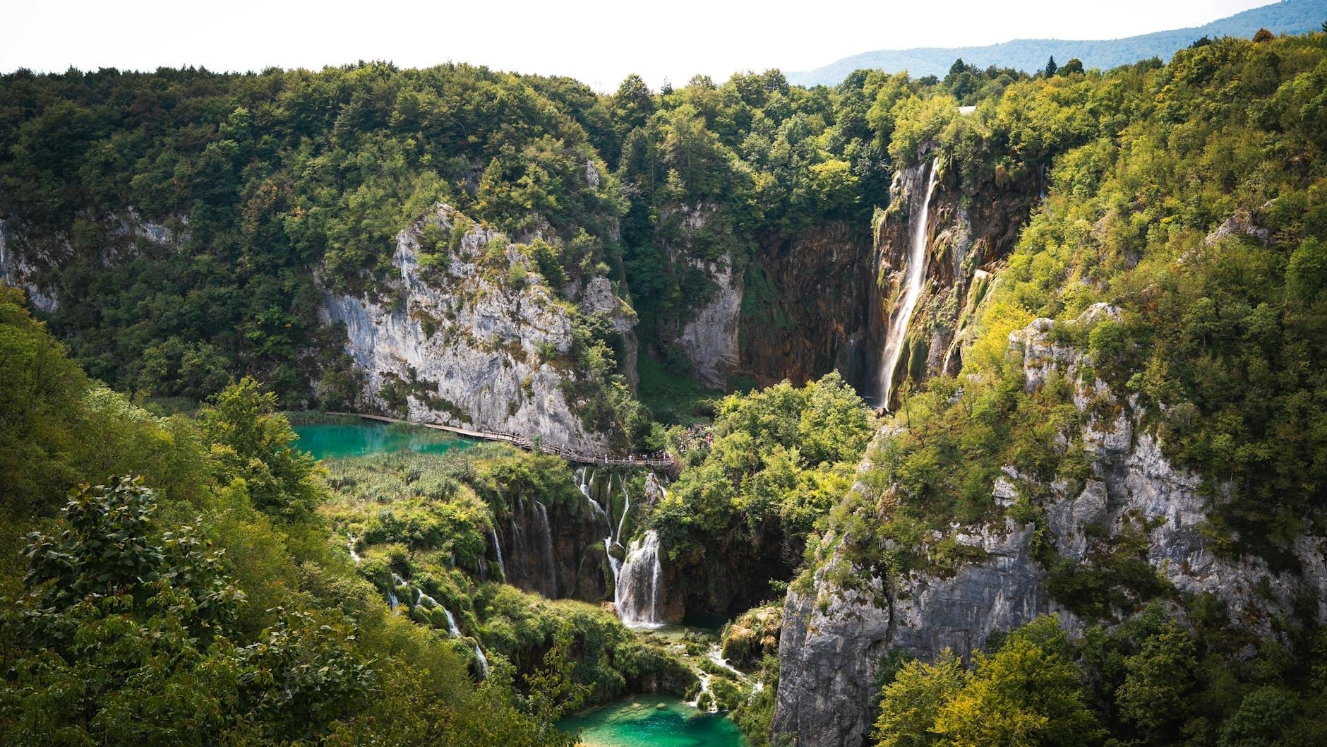 Vista panorâmica dos Lagos de Plitvice com suas piscinas esmeralda, cachoeiras dramáticas e falésias de calcário cobertas por floresta exuberante, captadas com uma luz natural deslumbrante.