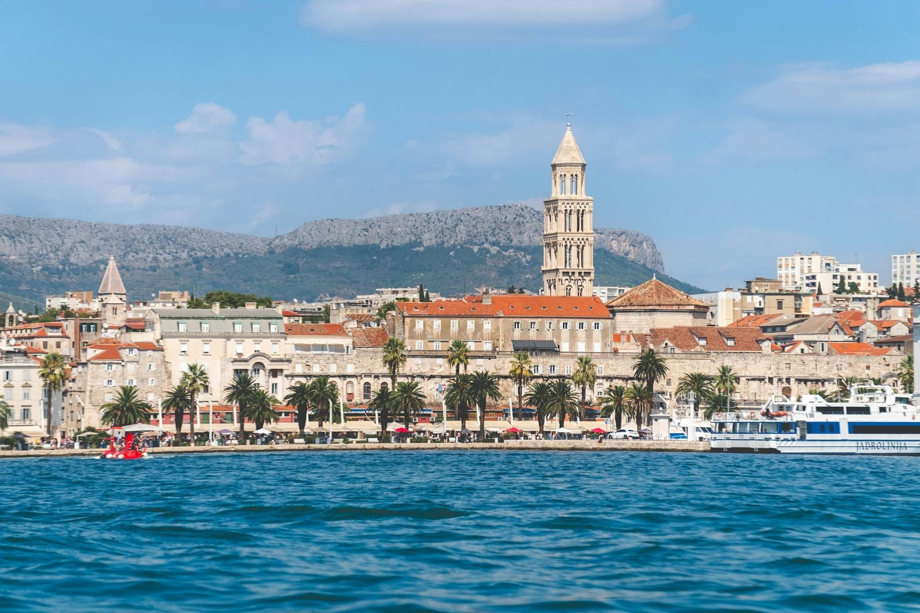 Vista panorâmica do calçadão de Split com palmeiras, barcos, o mar Adriático e uma torre sineira histórica ao fundo, com montanhas e céu azul intenso.