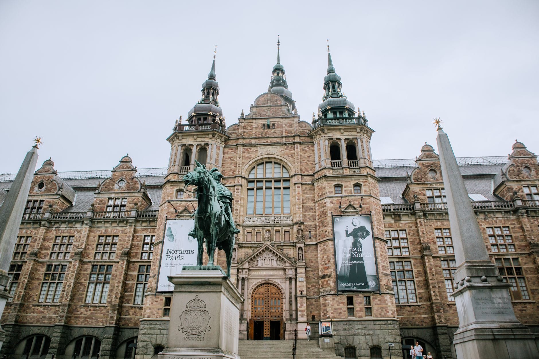 ด้านหน้าอาคารและหอคอยอันวิจิตรของ Nordic Museum ในสตอกโฮล์ม พร้อมรูปปั้นและเสาหินโอเบลิสก์ด้านหน้าในวันที่มีเมฆมาก