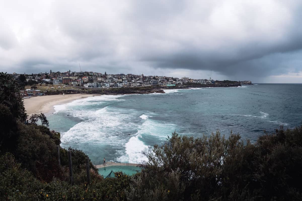 Vista panorámica de un espectacular litoral rocoso con olas en Bondi Beach y el barrio de Bondi al fondo bajo un cielo nublado.
