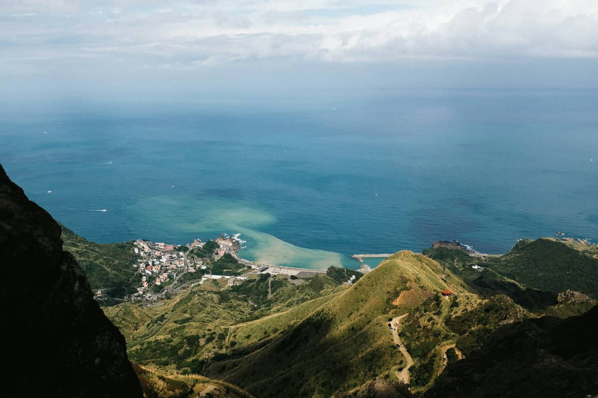 Weitwinkelperspektive auf üppig begrünte Küstenberge, die auf ein kleines Städtchen am Meer und den blauen Ozean treffen – ein Vorgeschmack auf die Ausflugsziele rund um Taipeh.