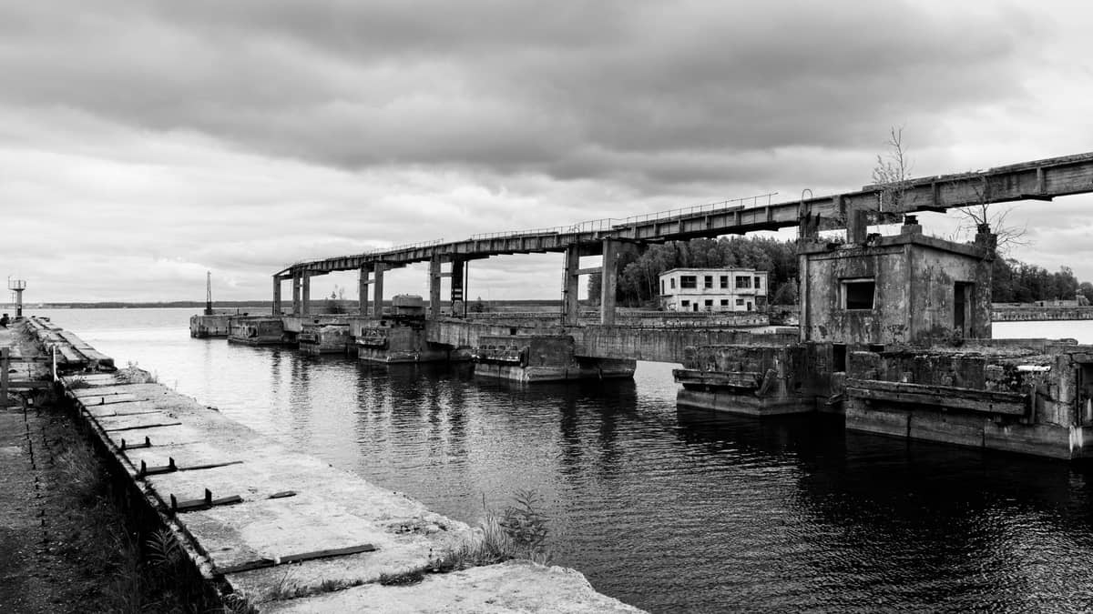 Foto en blanco y negro de un sitio industrial abandonado de la era soviética con estructuras de concreto en deterioro sobre el agua y un cielo sombrío.