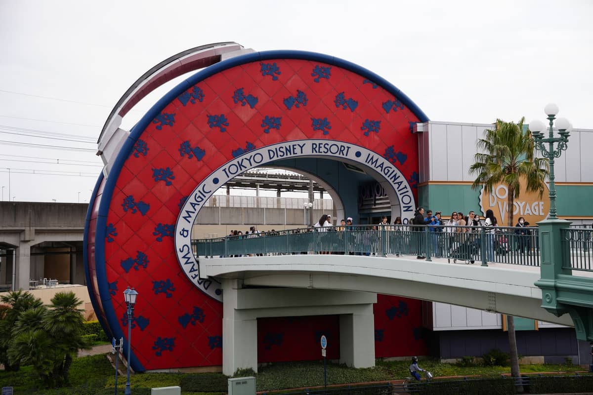 Entrance to Tokyo Disney Resort with a distinctive red and blue circular structure and crowds of people walking over a bridge.