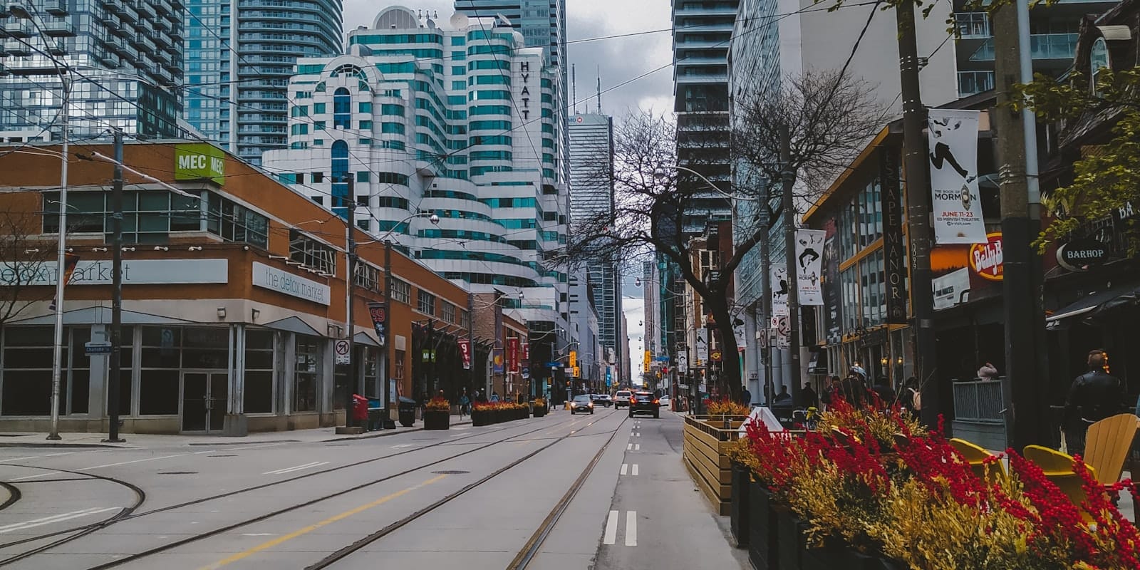 Vue d'une rue animée du centre-ville de Toronto avec des immeubles modernes, des commerces de détail et des rails de tramway par une journée nuageuse.