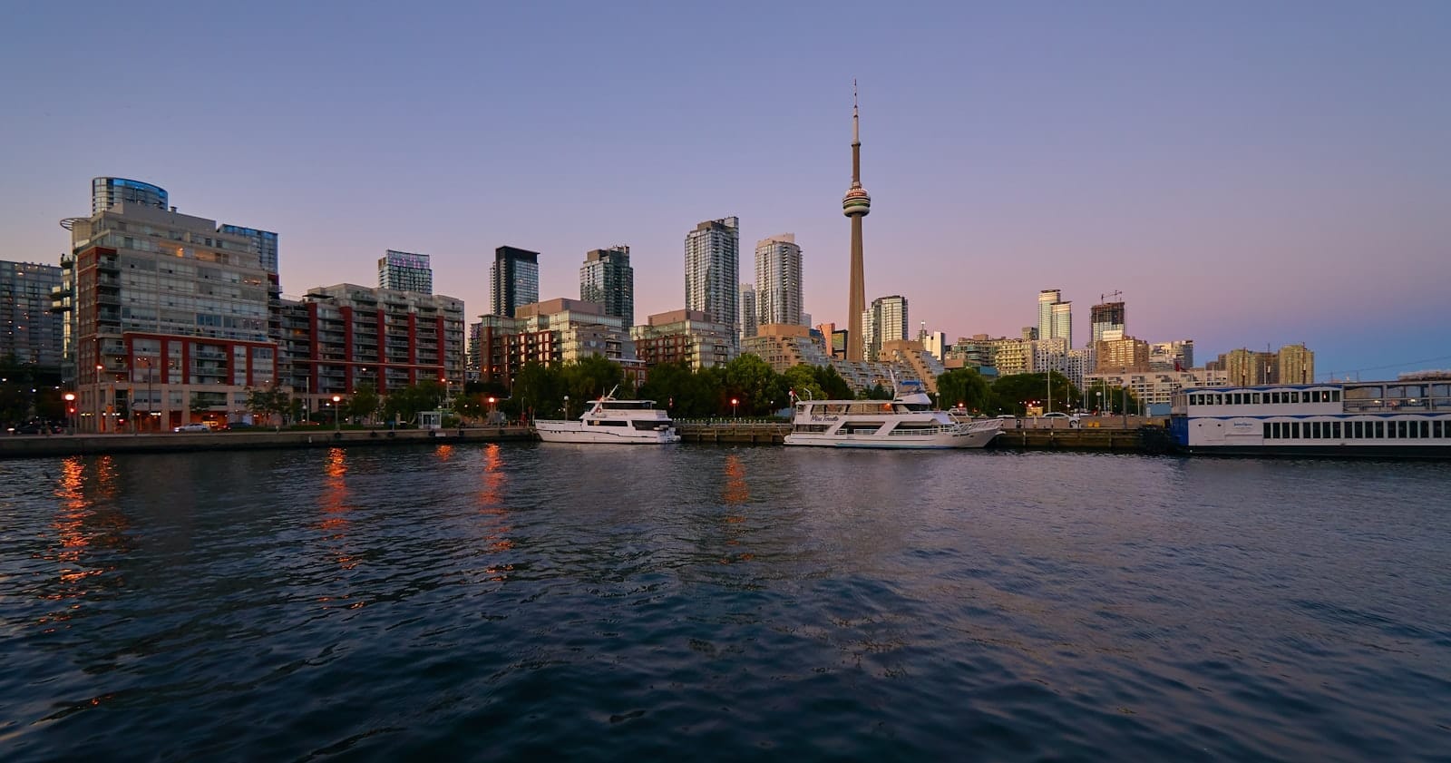 Front de mer de Toronto avec des bateaux amarrés le long du port, horizon moderne et la CN Tower en arrière-plan au coucher du soleil.