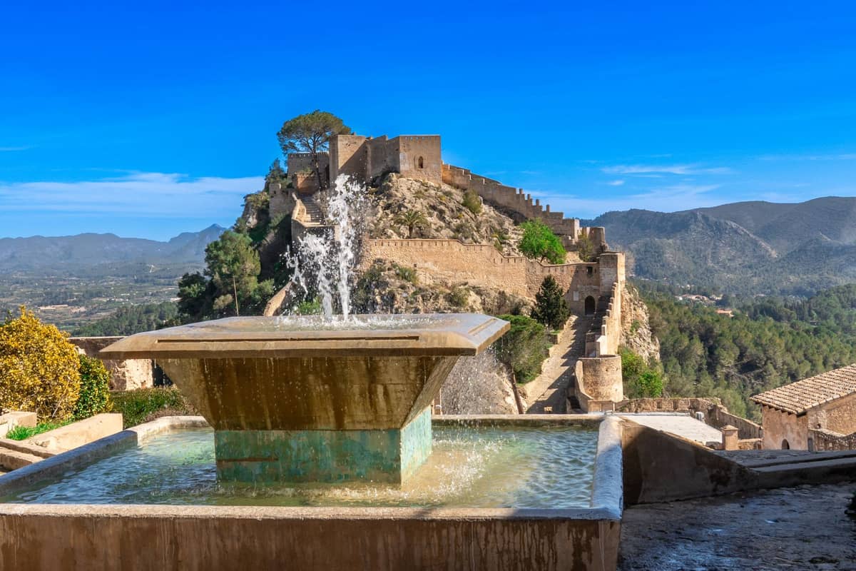 Vista del castello storico di Xàtiva su una collina, con mura in pietra, torri di avvistamento e una fontana in primo piano, circondata da montagne boscose.