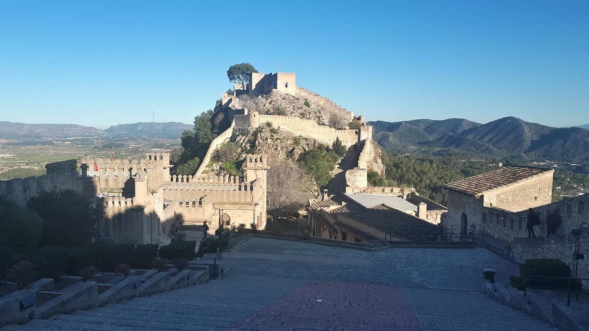 Weitblick auf die Burg Xàtiva auf einem steilen Hügel mit Mauern, Türmen und Wehranlagen, mit Blick auf grüne Täler und ferne Berge unter blauem Himmel.