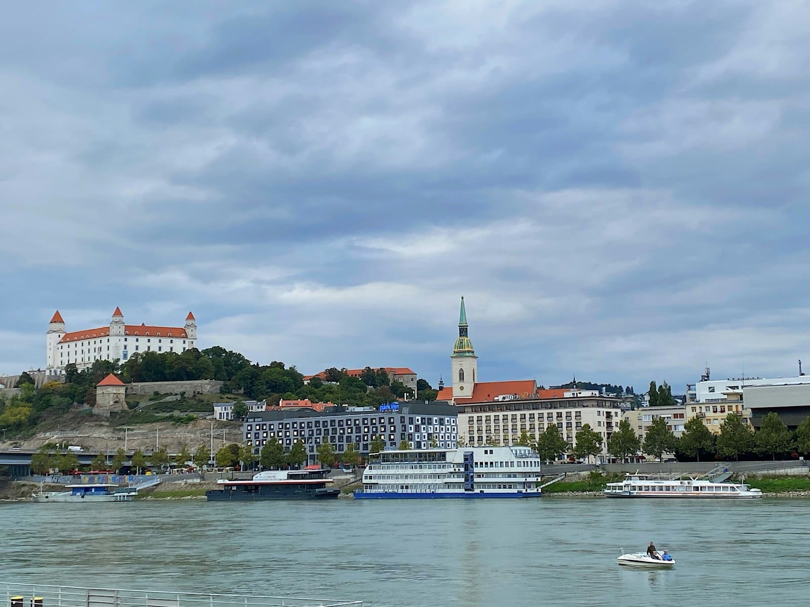 Castillo de Bratislava y Catedral de San Martín con el río Danubio y barcos en primer plano bajo un cielo nublado.
