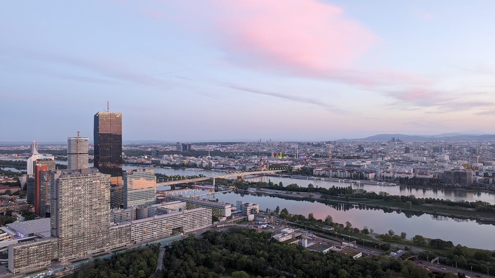 Vista aérea amplia de Viena que muestra el río Danubio, modernos edificios de gran altura, puentes y el paisaje urbano lejano bajo un cielo rosado y azul.