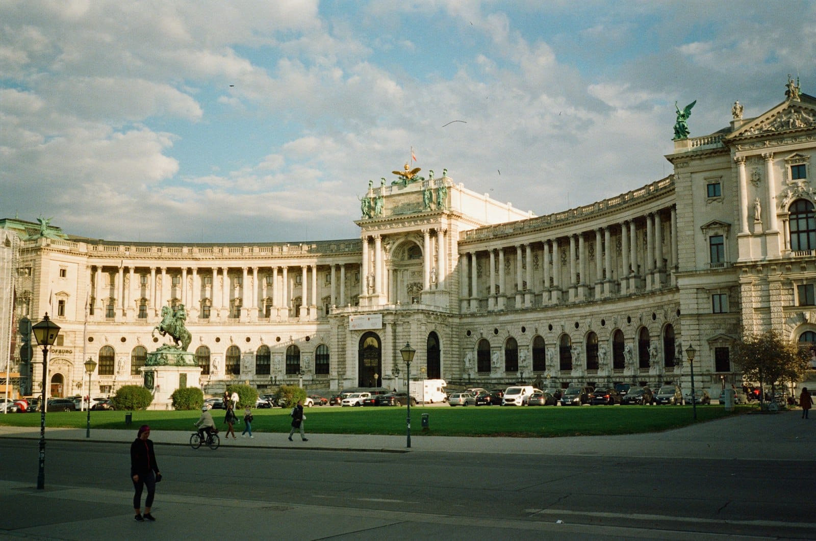 Weiter Blick auf die Wiener Hofburg mit Besuchern und Radfahrern auf dem Platz unter einem dramatisch bewölkten Himmel – das ideale Tor für Tagesausflüge.
