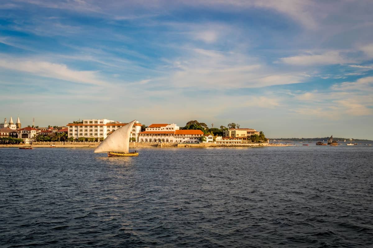 Dhow tradizionale che naviga vicino al lungomare di Stone Town con edifici storici visibili lungo la costa sotto un cielo azzurro.